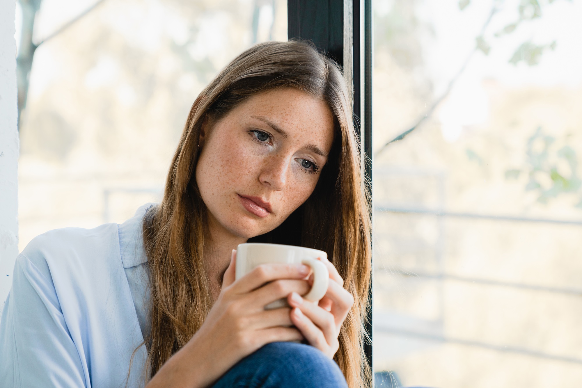 Sad young woman, the victim of abuse, thinking about trauma at home by the window alone.